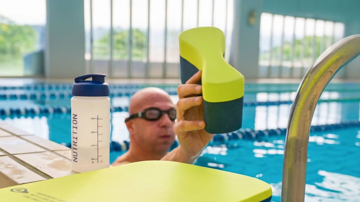 Un nuotatore in piscina solleva un pull buoy giallo, con borraccia e tavoletta appoggiate sul bordo vasca