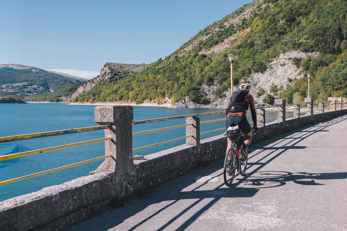 Ciclista con la bici carica di borse pedala sulla strada che costeggia un lago tra le colline