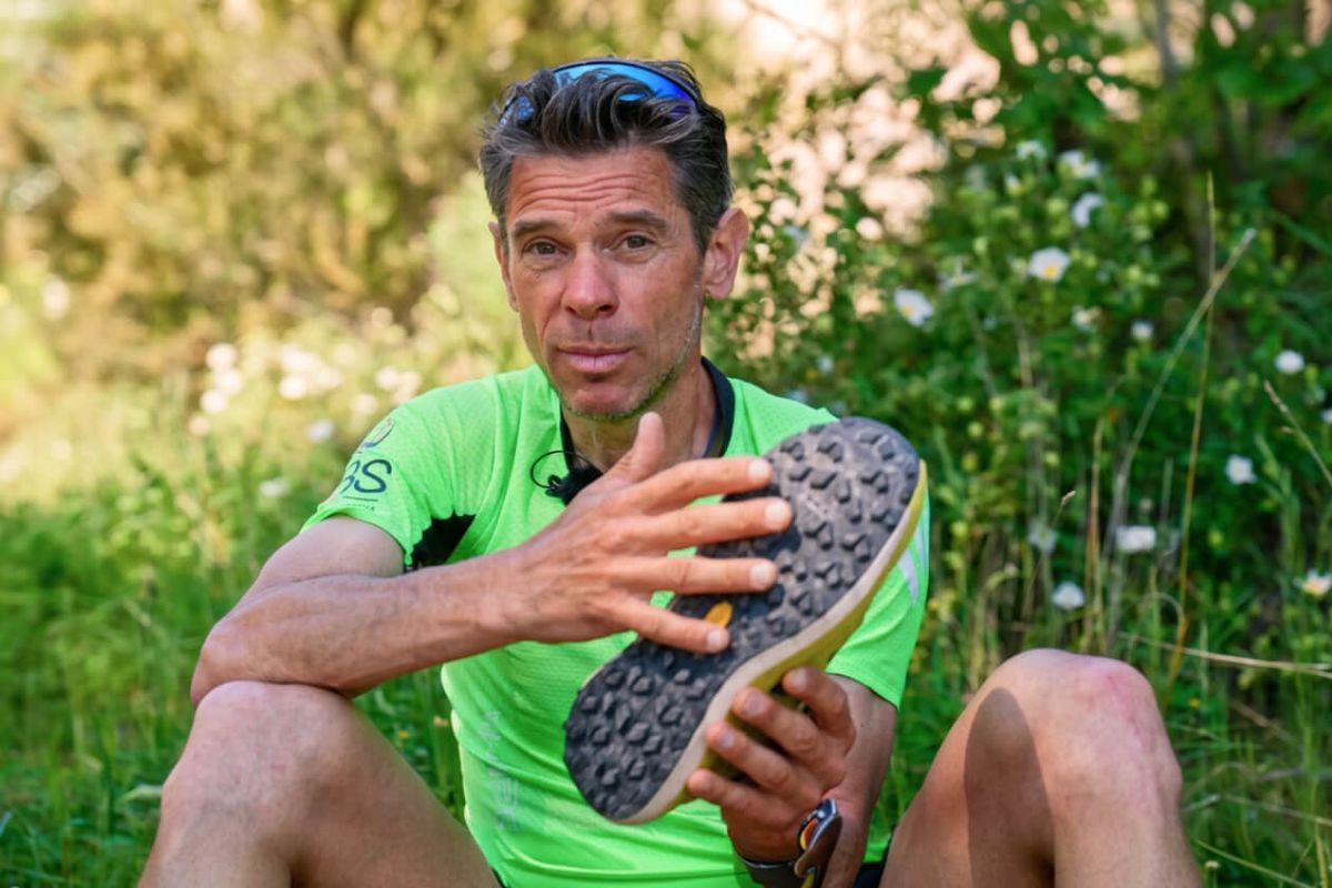 Marco De Gasperi, seduto su un prato, mostra la suola tassellata di una scarpa da trail running