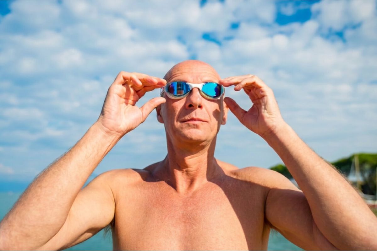 Luca Sacchi si sistema gli occhialini da nuoto a specchio, a torso nudo in riva al mare sotto un cielo con nuvole sparse