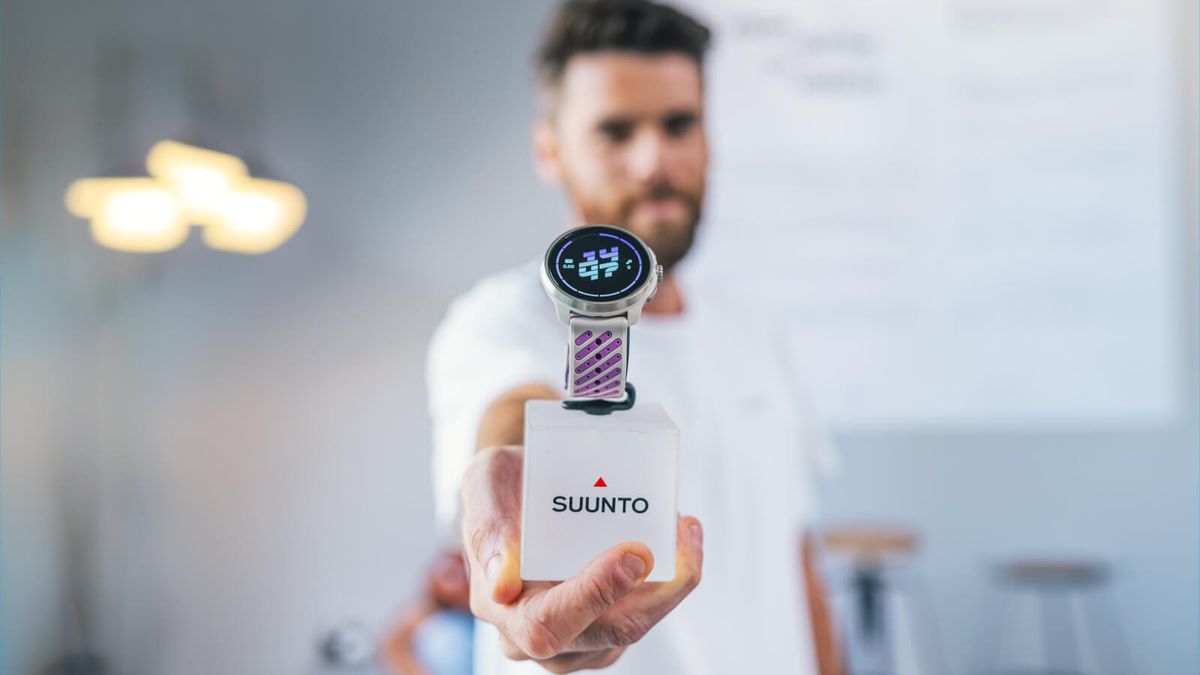 Un uomo mostra verso l'obiettivo un orologio Suunto di ultima generazione con il cinturino viola, appoggiato su un cubo con il logo Suunto