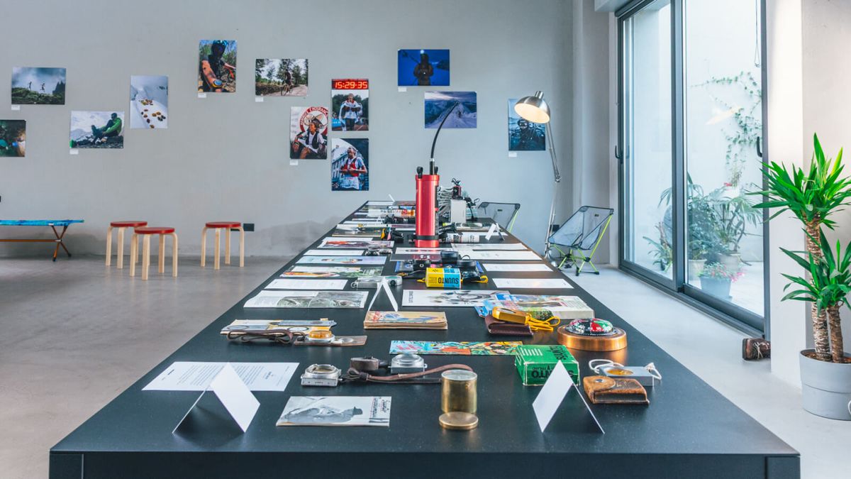 La sala della mostra Suunto From Depths to Heights a Milano: un lungo tavolo nero con bussole, orologi e cataloghi d'archivio, fotografie alle pareti e sgabelli in legno
