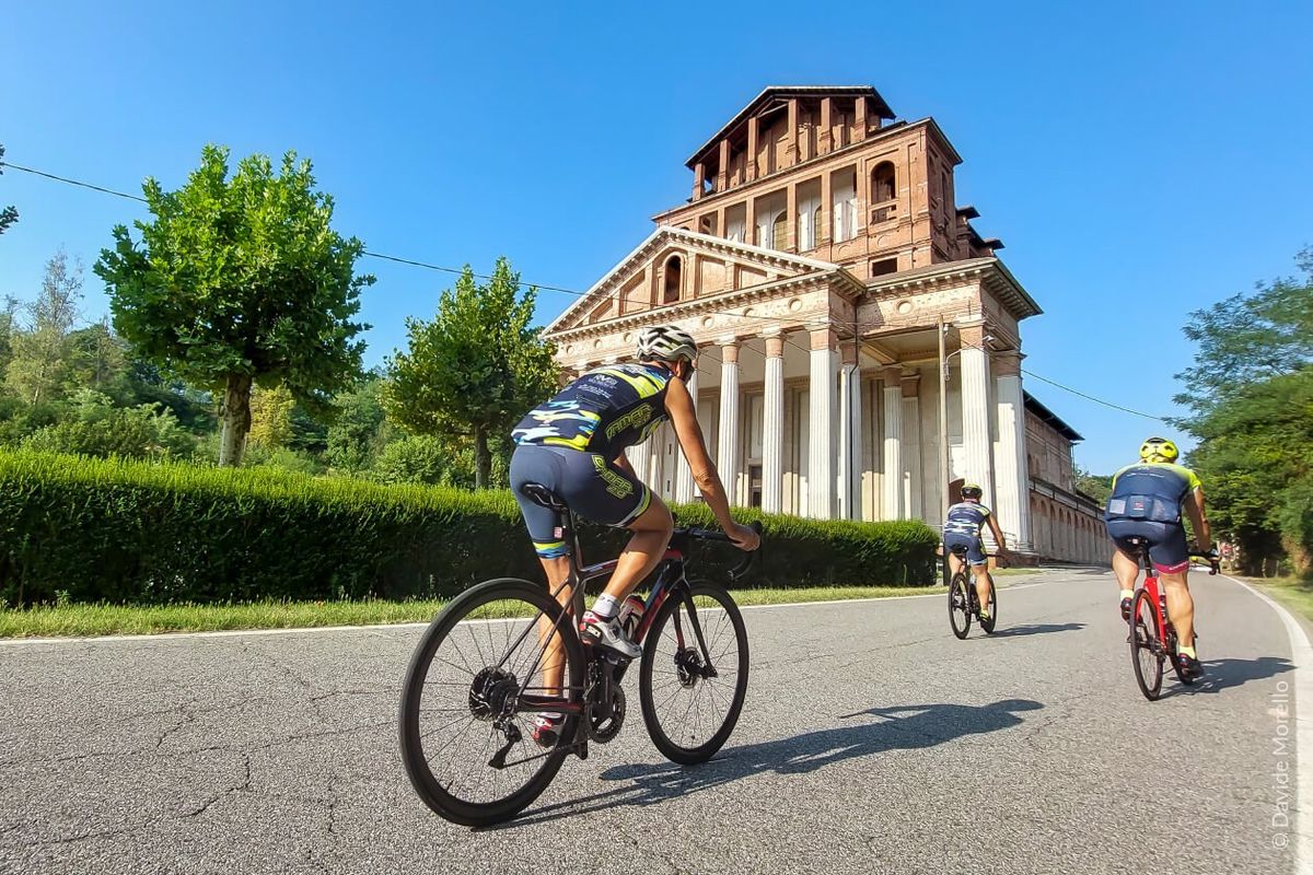 Tre ciclisti su strada pedalano davanti a un edificio storico neoclassico in mattoni