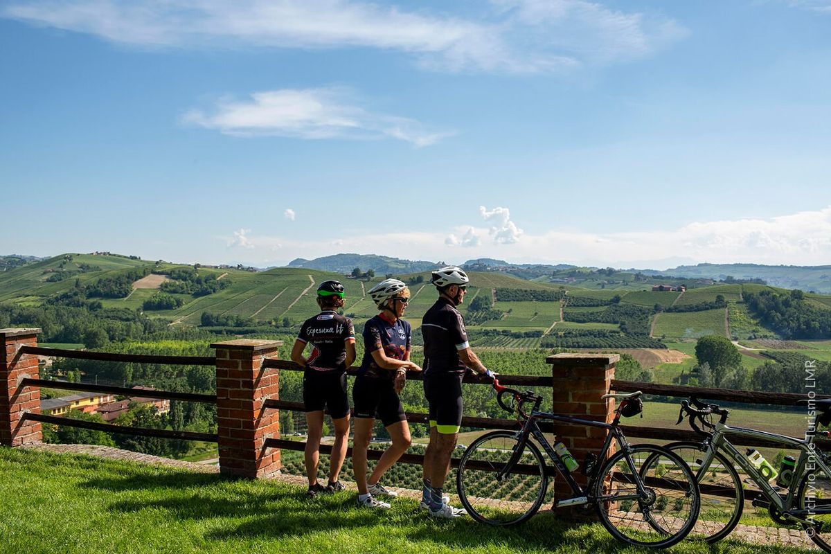 Tre ciclisti su bici da corsa si fermano a un belvedere con vista sulle colline vitivinicole delle Langhe
