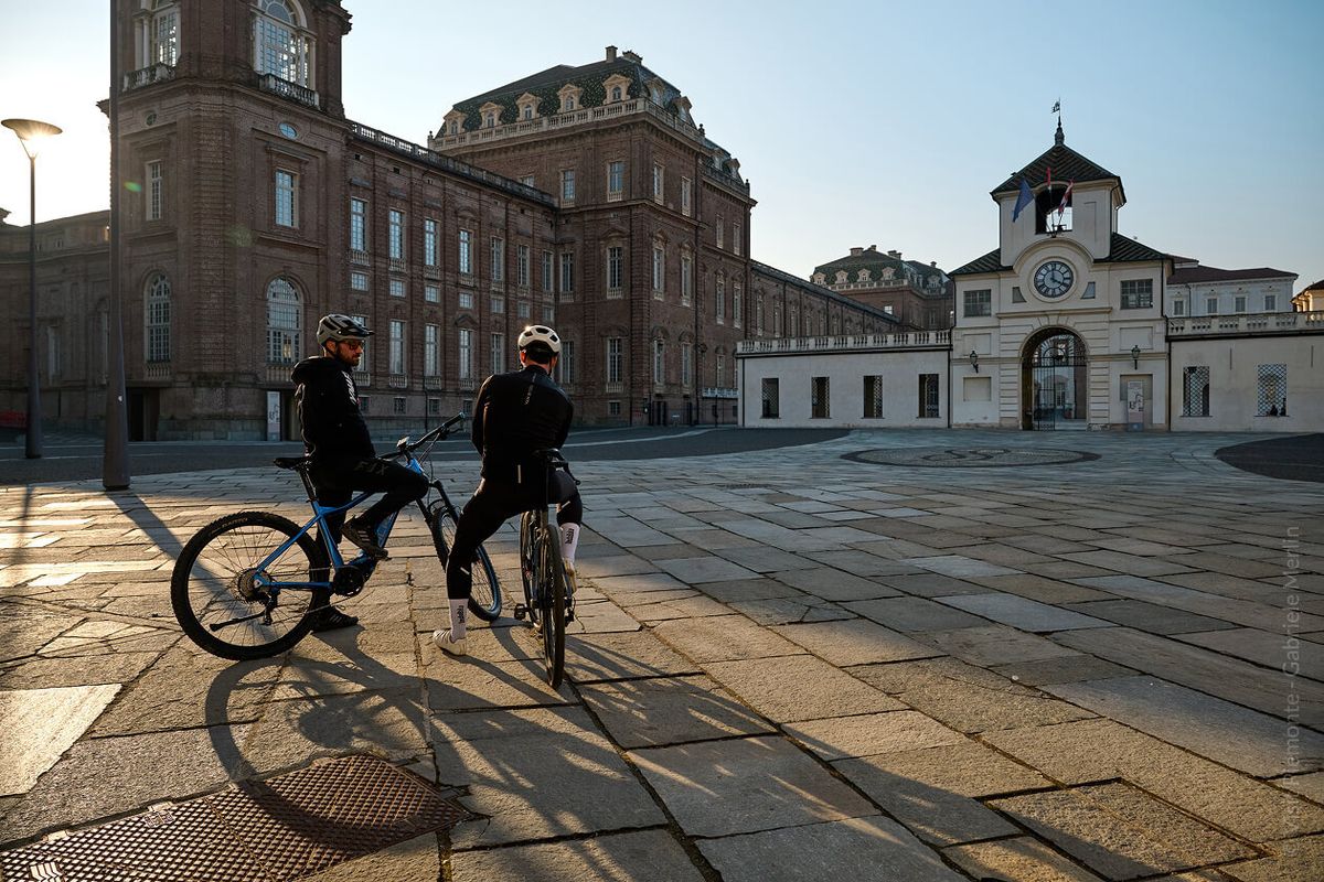 Due ciclisti in sosta nella piazza della Reggia di Venaria Reale all'alba, davanti alla facciata barocca e alla torre dell'orologio