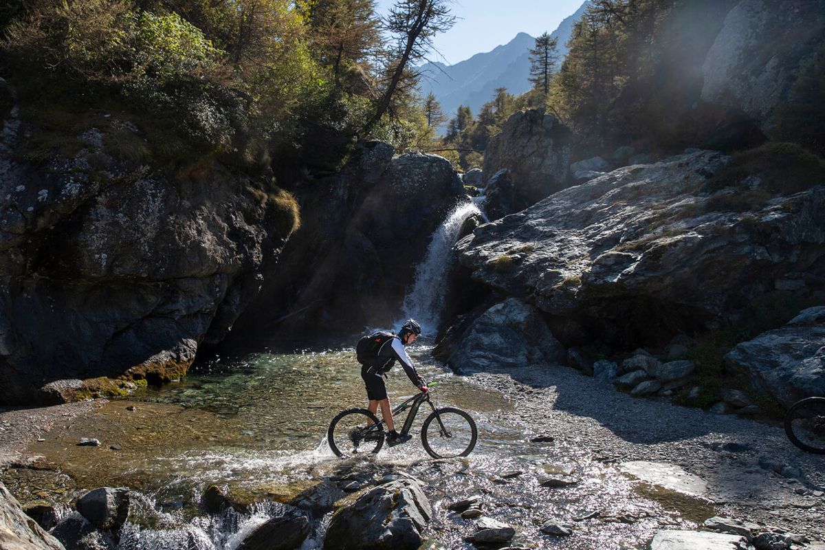 Biker in mountain bike attraversa un torrente alpino accanto a una cascata tra le rocce