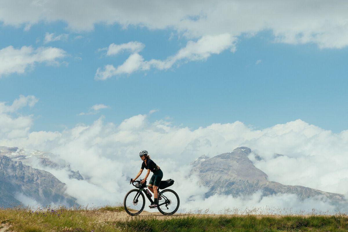 Ciclista in gravel pedala in quota su un crinale erboso sopra un mare di nuvole, con le cime alpine sullo sfondo