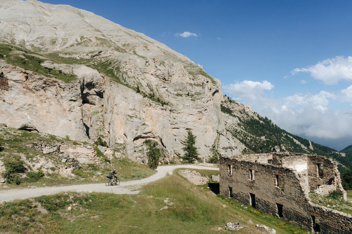 Ciclista solitario su una strada sterrata di alta quota ai piedi di una parete rocciosa, accanto ai ruderi di un edificio in pietra