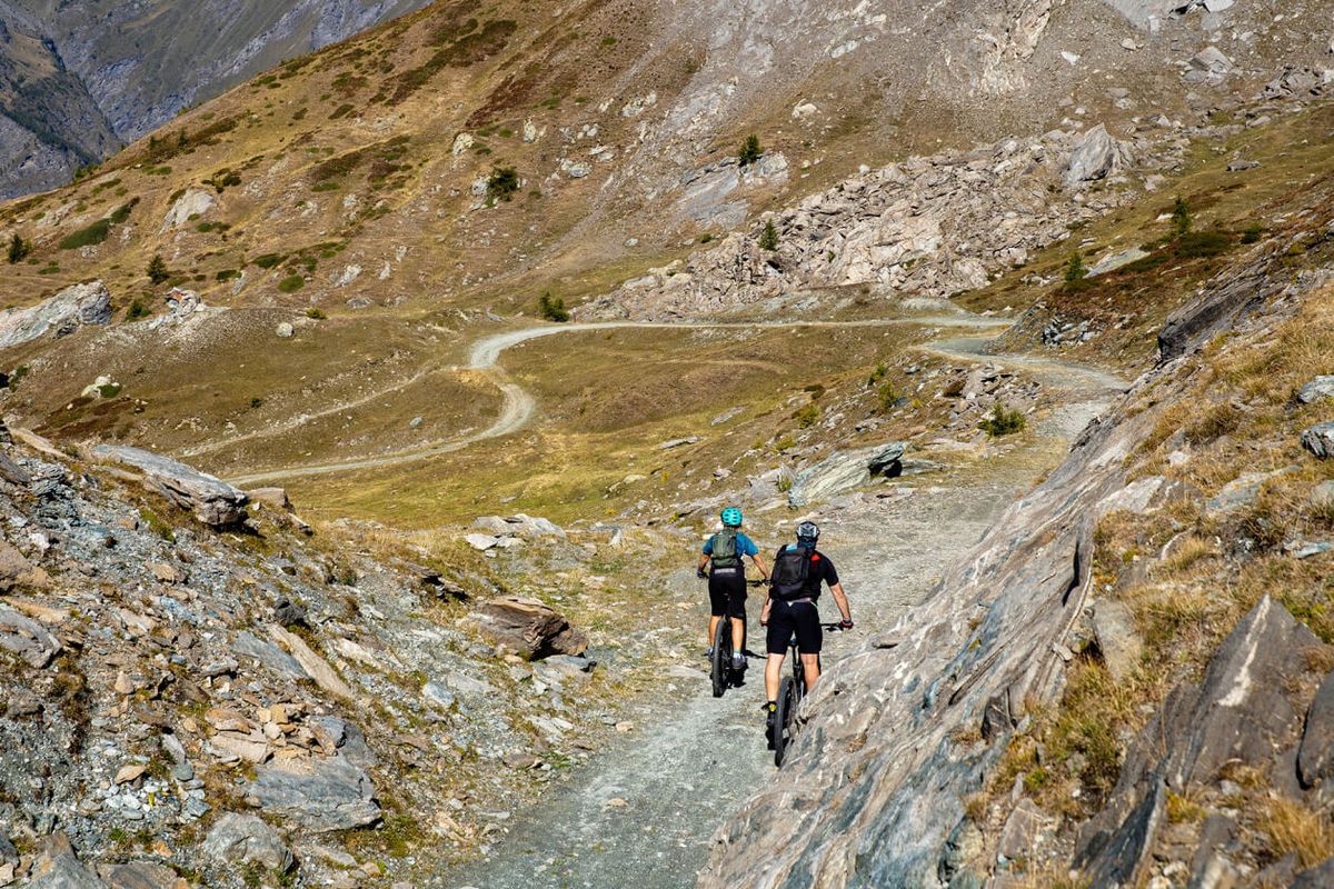 Due biker di spalle percorrono un sentiero sterrato tra i pendii rocciosi di alta quota